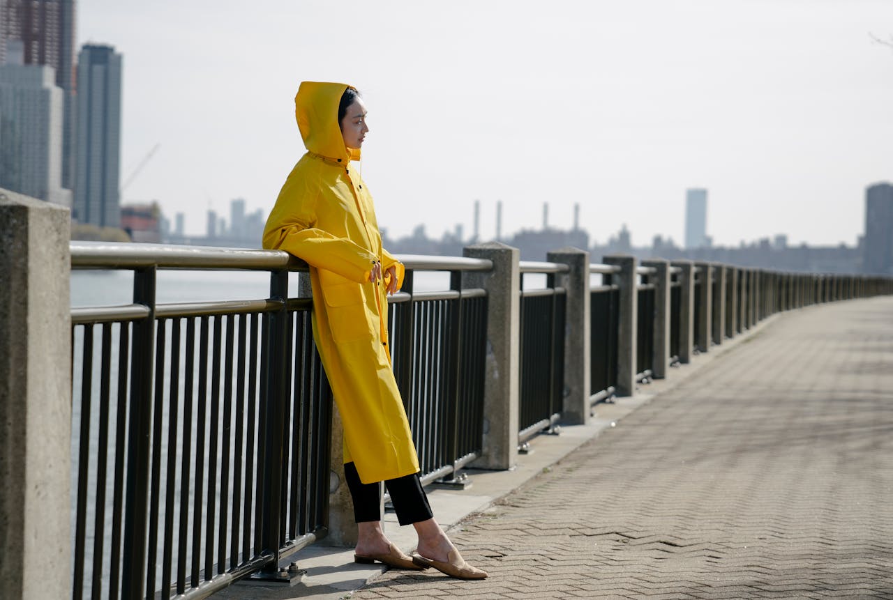 Woman in a yellow raincoat on a city waterfront, leaning on railing.
