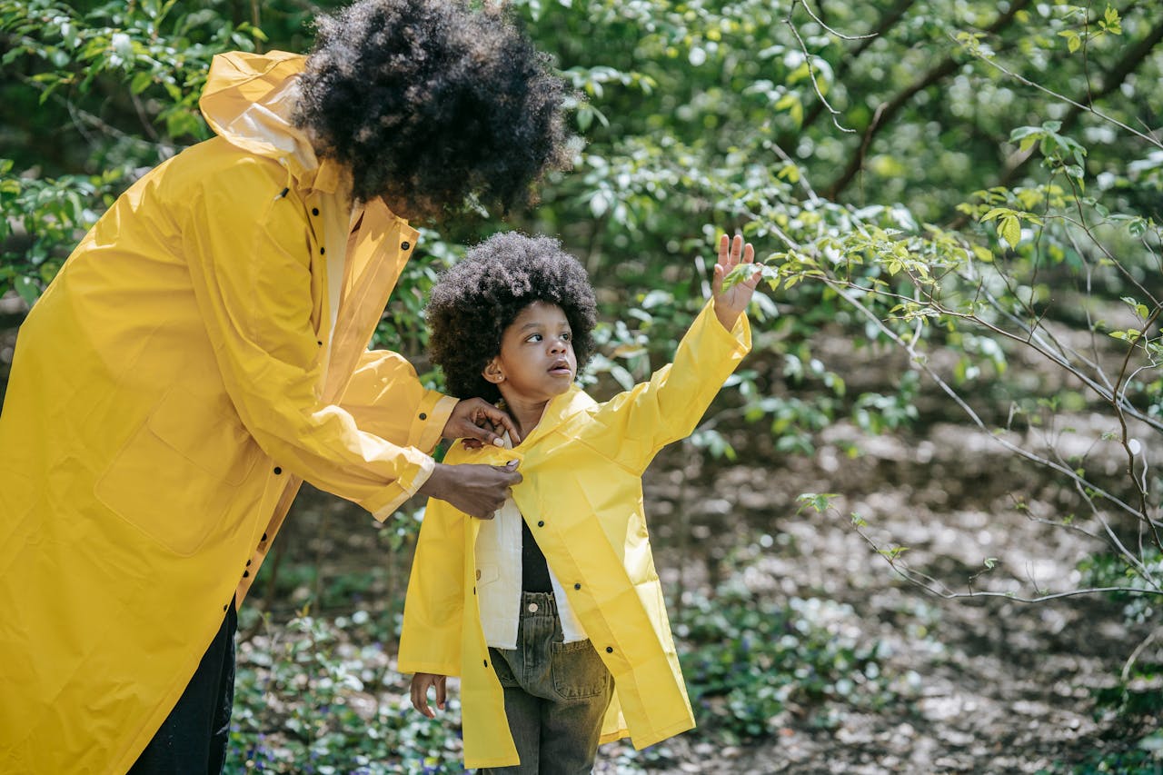 A mother helps her child button a yellow raincoat while exploring a lush green forest.